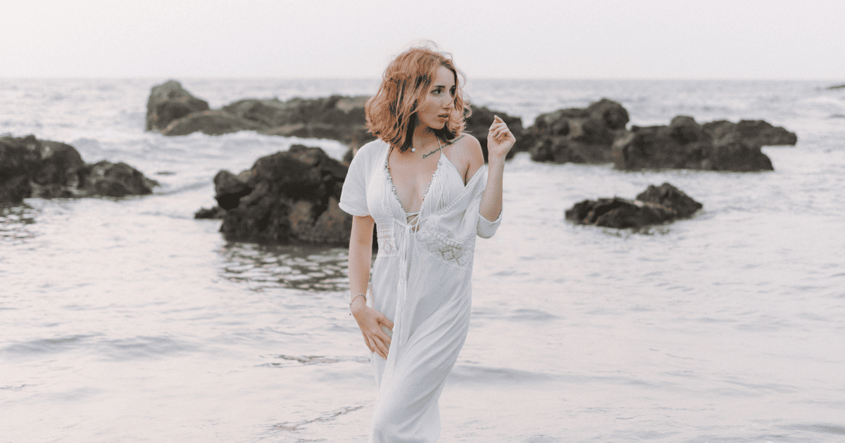 redheaded woman in a white dress standing in water at the beach | How To Become Magnetic To Your Dreams | The Aligned Life Manifestation Blog